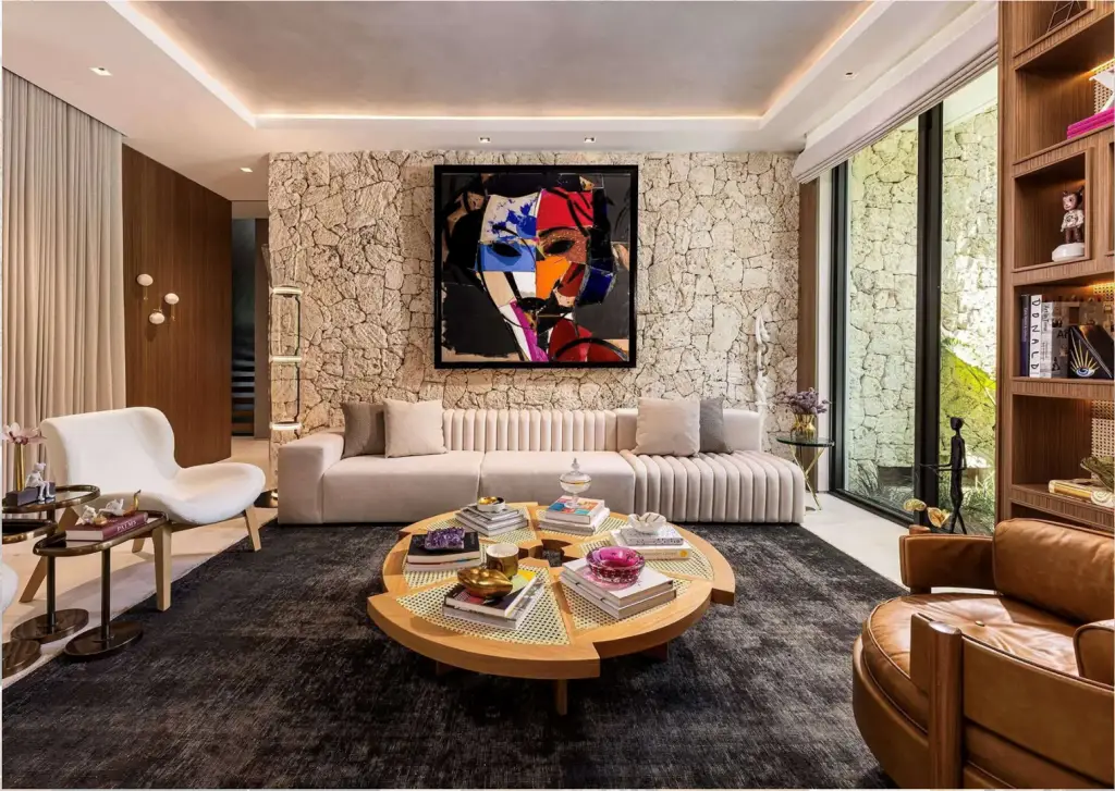 Contemporary living room with a textured stone accent wall, abstract artwork, neutral-toned seating, a round wood coffee table, and built-in shelving.