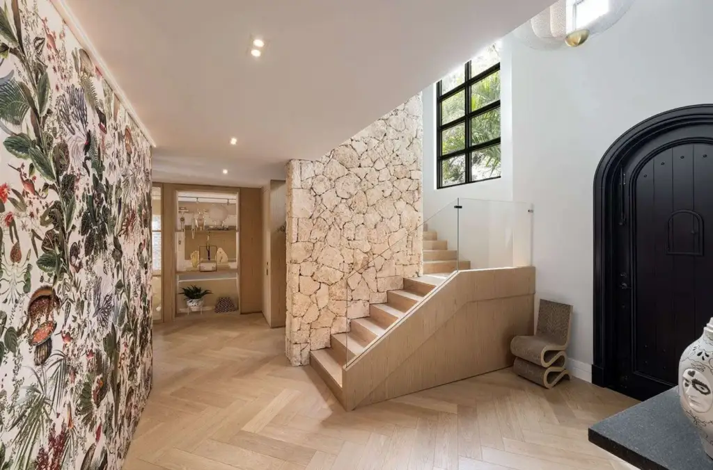 Bright entryway with coral stone staircase, arched black door, large modern window, and nature-inspired wallpaper in a historic Coral Gables home.