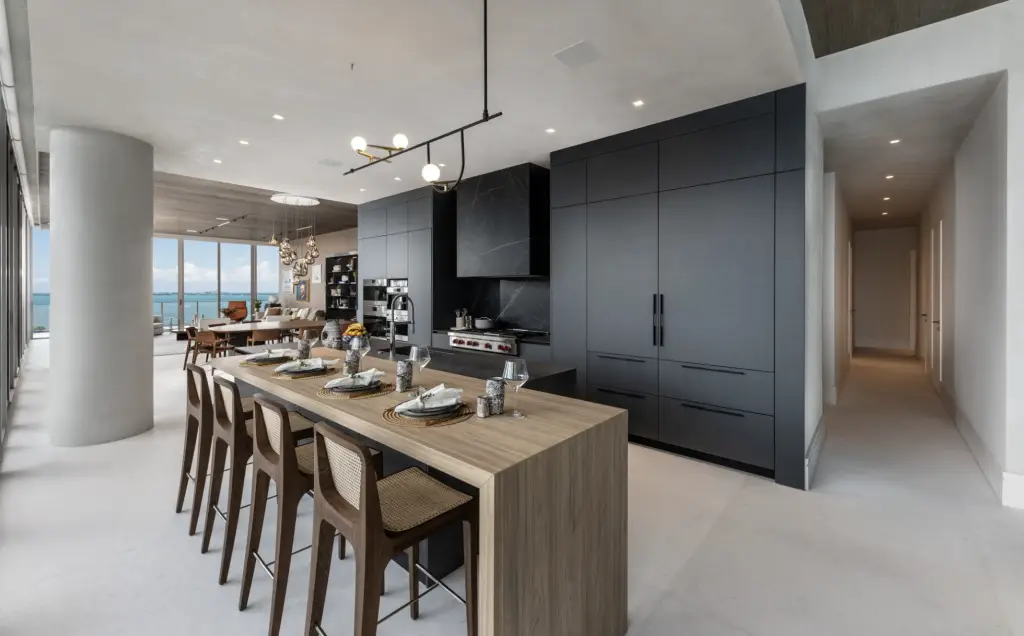 Modern open-concept kitchen with matte black cabinetry, wood island with bar seating, minimalist lighting, and floor-to-ceiling oceanfront views in a Coconut Grove high-rise.