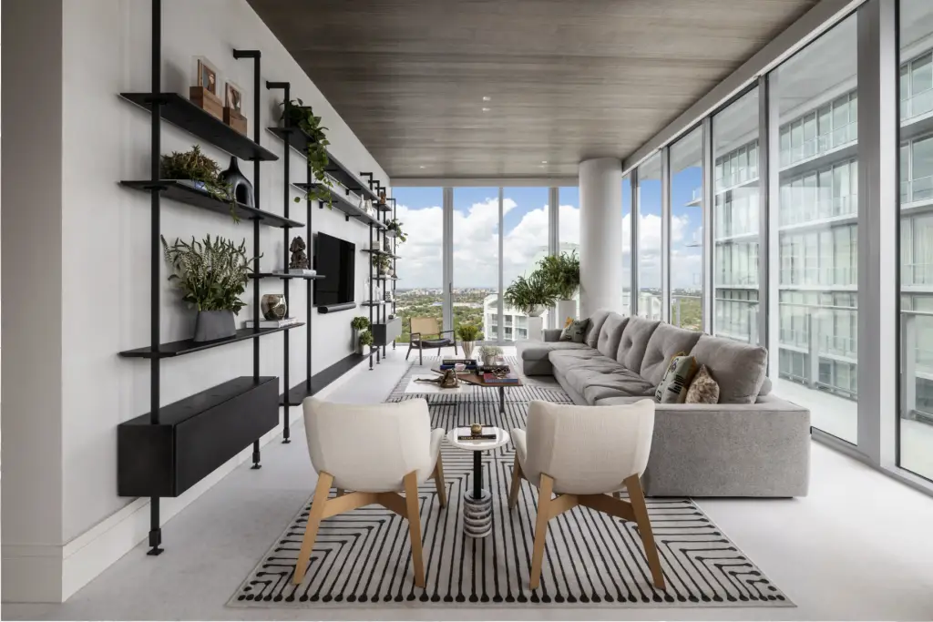 Contemporary high-rise living room in Coconut Grove featuring floor-to-ceiling windows, a gray sectional sofa, black open shelving with greenery, and a striped area rug for a modern, airy ambiance.